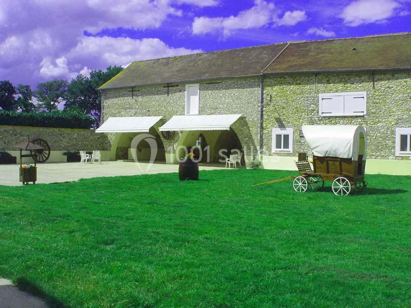 Cour d'une ferme en pierre avec pelouse, charrette en bois, tonneau fleuri et ciel partiellement nuageux.