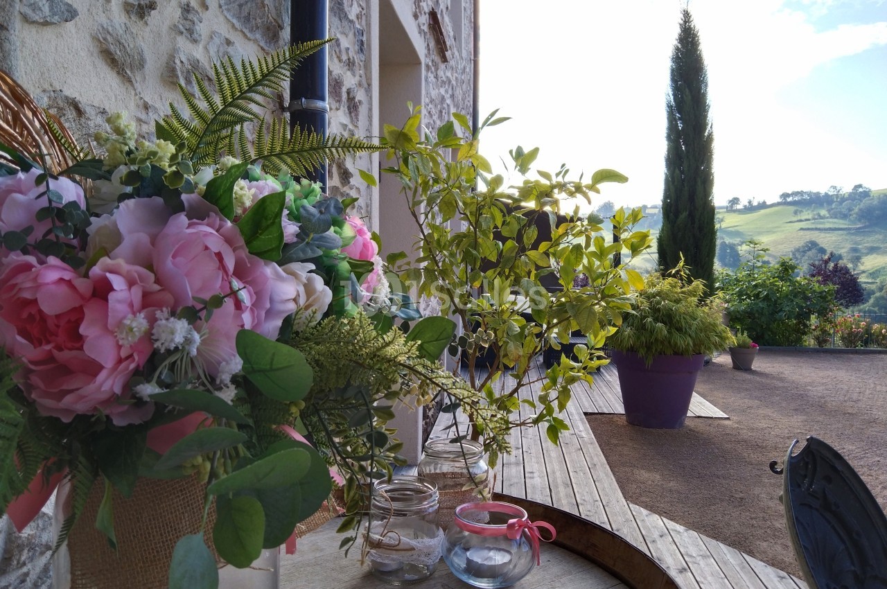 Bouquet de fleurs roses et feuillages sur une table en bois, avec vue sur un jardin et un paysage vallonné en arrière-plan.