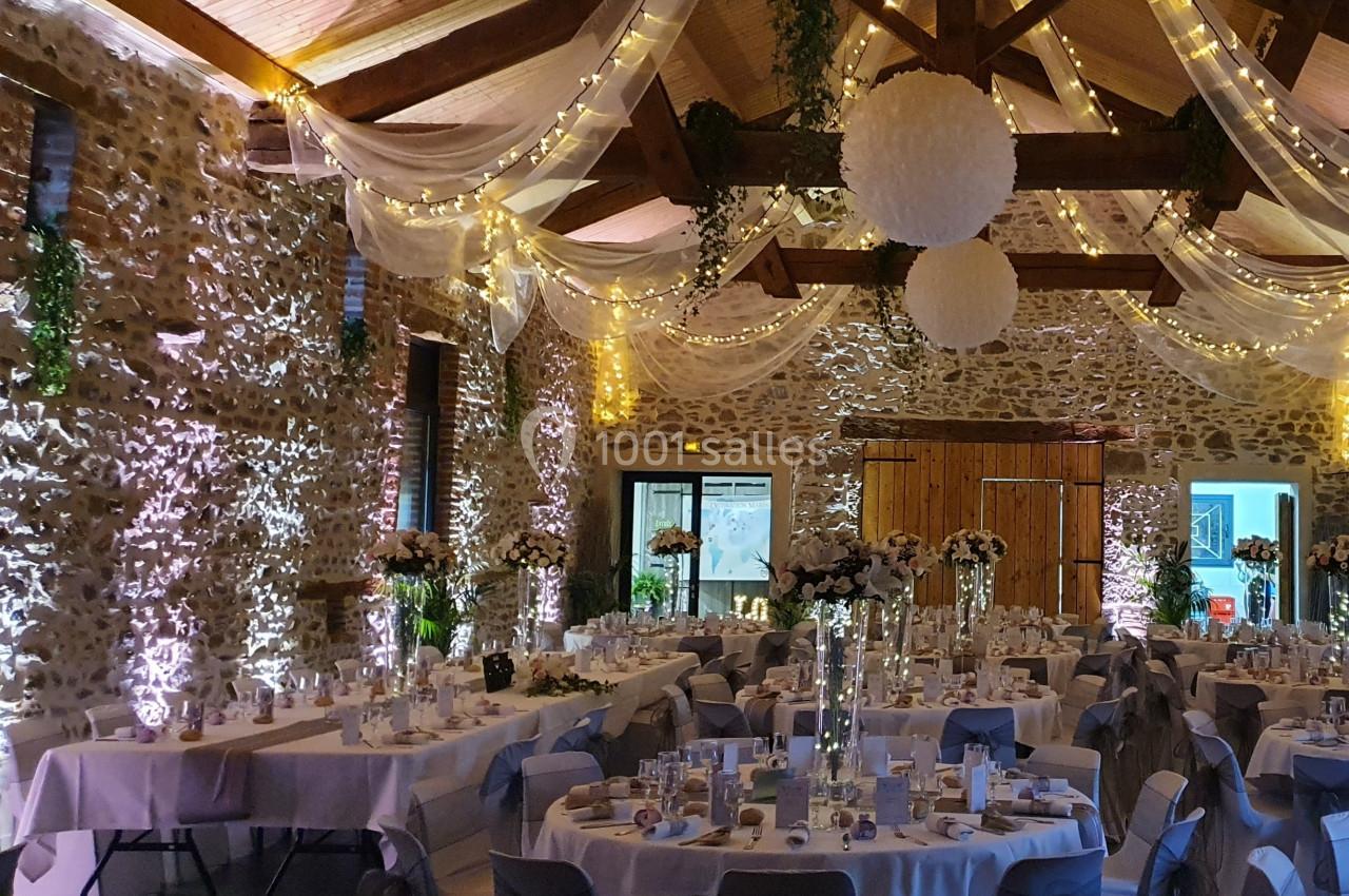 Salle de réception décorée avec des guirlandes lumineuses, des tables rondes dressées et des suspensions blanches.