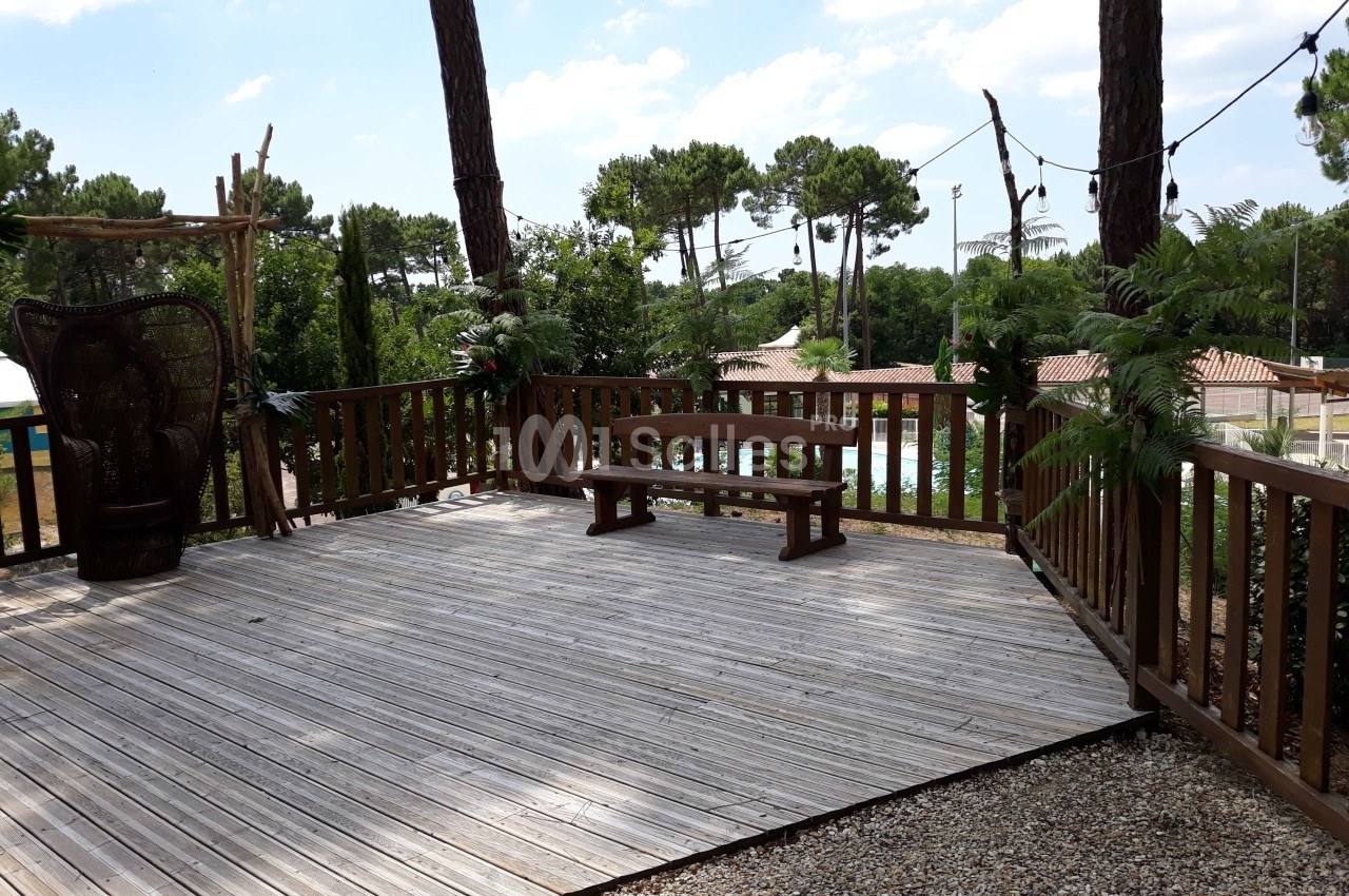 Terrasse en bois entourée de végétation, avec une table et un banc, sous un ciel partiellement nuageux.