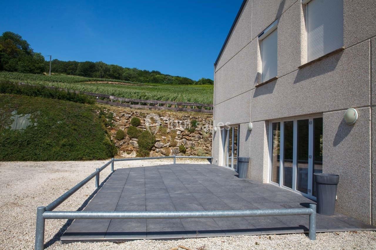 Terrasse en dalles grises devant un bâtiment moderne, entourée de barrières, avec un paysage de vignes en arrière-plan.