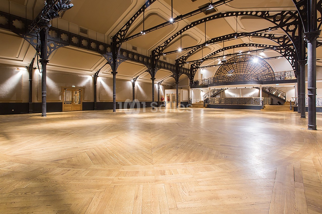 Grande salle vide avec parquet en bois clair, structure métallique apparente et éclairage suspendu.