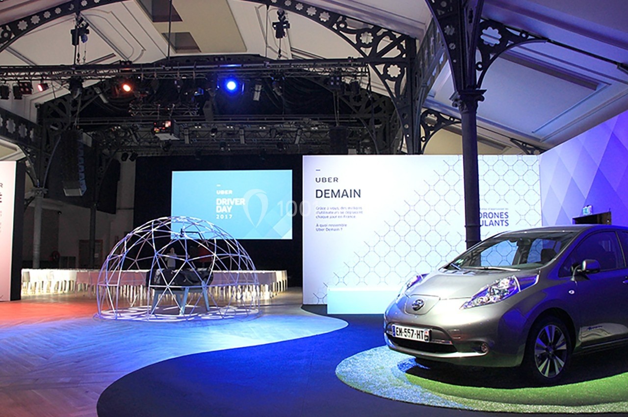 Salle d'événement avec une voiture électrique exposée, une structure géodésique et des écrans affichant des présentations.