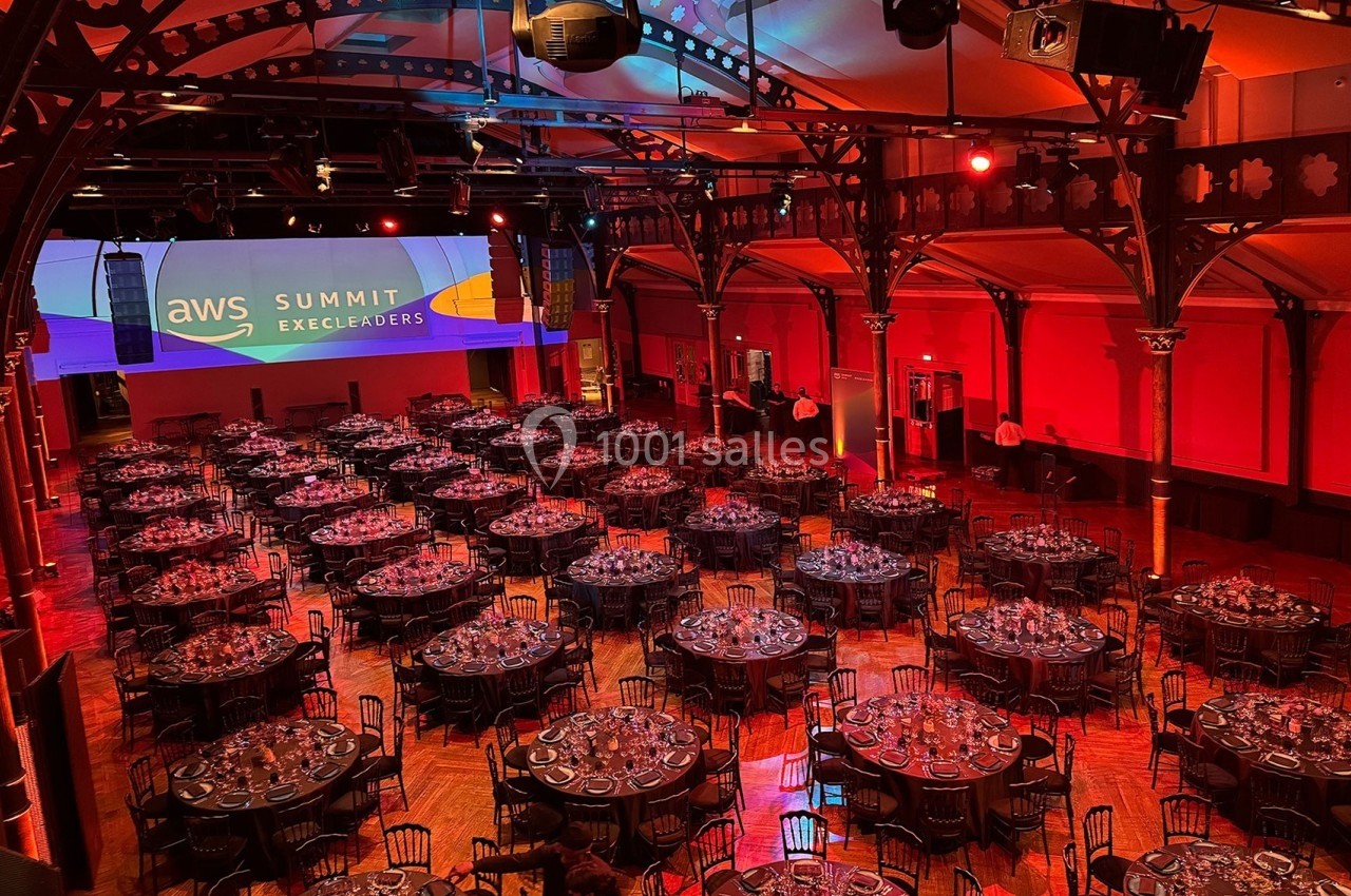 Salle de réception avec tables rondes dressées pour un événement, éclairée par des lumières rouges et un écran projetant…