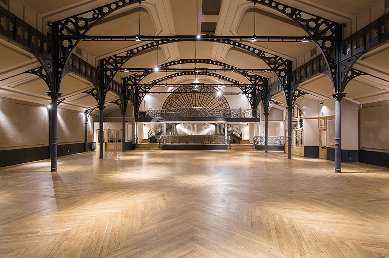 Grande salle vide avec parquet en bois, structure métallique apparente et plafond voûté éclairé par des spots.