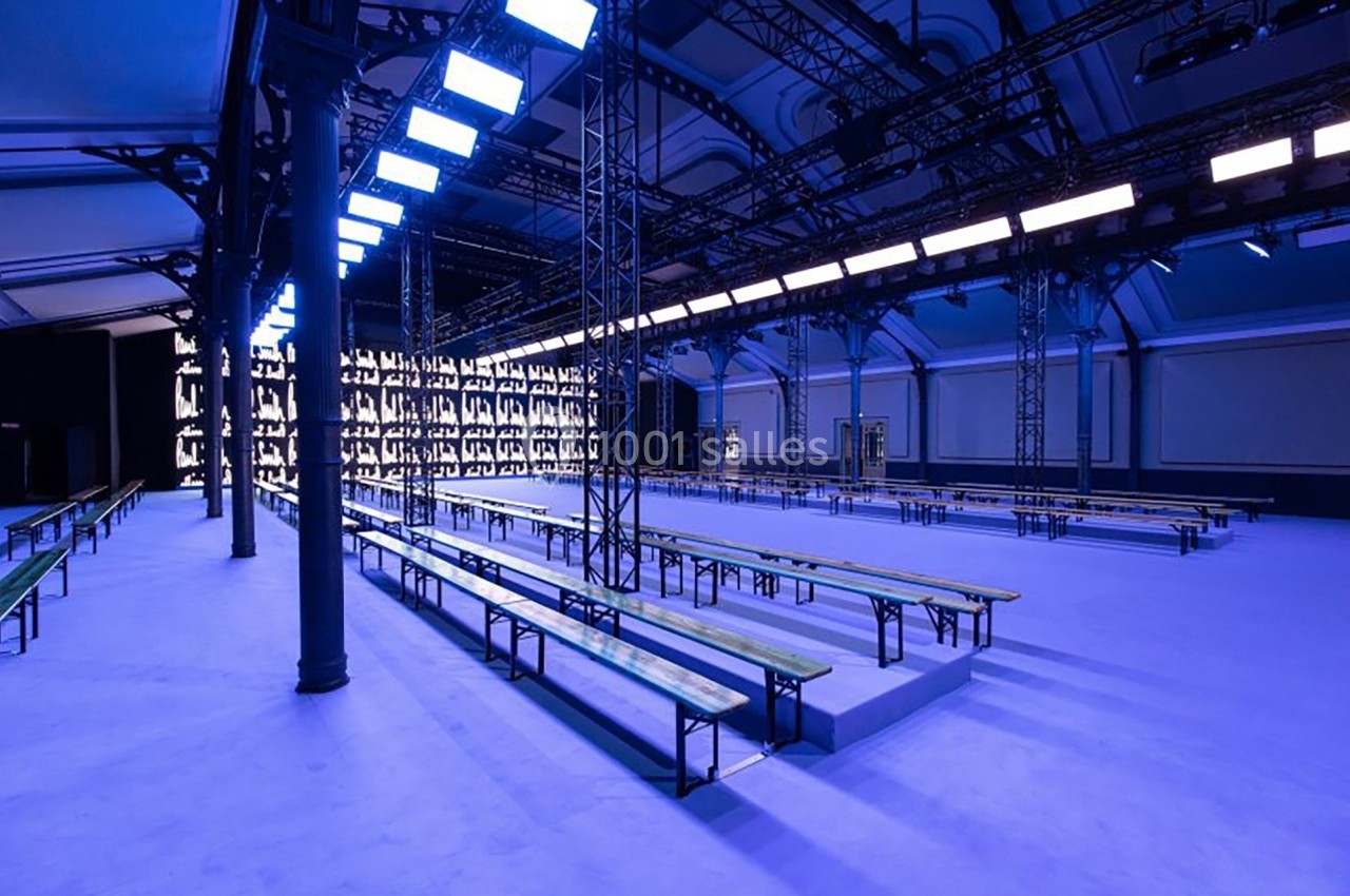 Salle vide avec bancs alignés, éclairage bleu et structure métallique, préparée pour un événement ou un défilé.