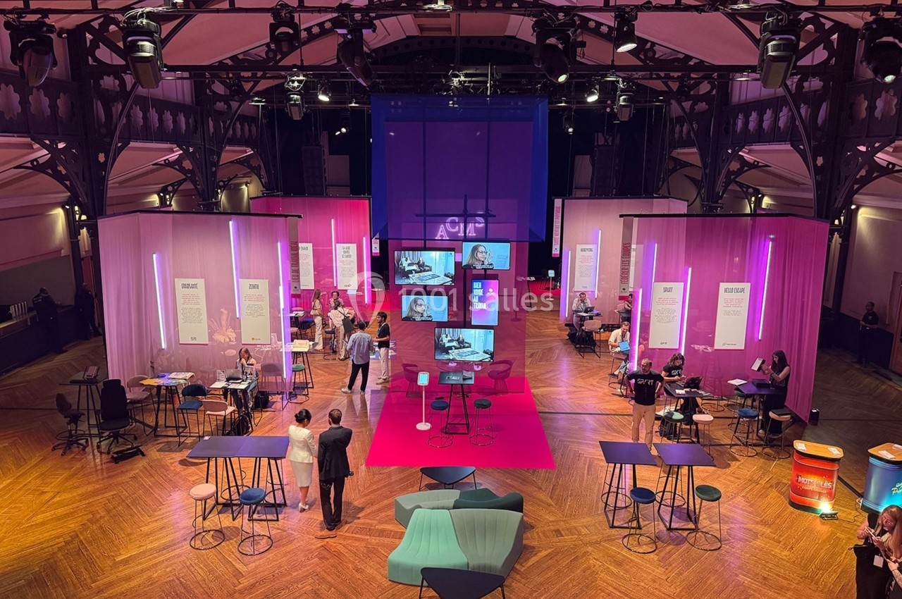 Salle événementielle avec éclairage coloré, stands d'exposition, tables hautes et visiteurs discutant.