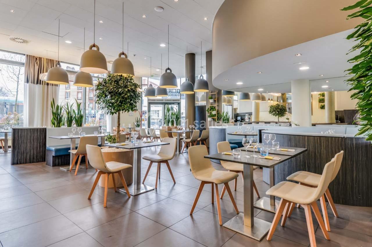 Salle de restaurant moderne avec tables dressées, chaises en bois clair, luminaires suspendus et plantes décoratives.