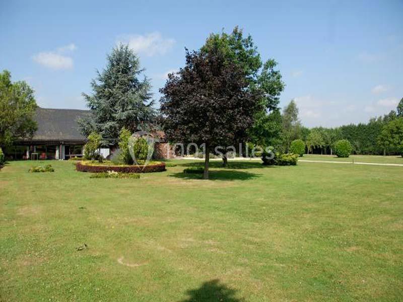 Pelouse verdoyante avec quelques arbres, arbustes et une maison en arrière-plan sous un ciel dégagé.