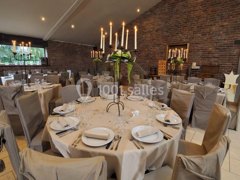 Salle de réception avec tables rondes dressées, nappes blanches, chandeliers et chaises couvertes de housses grises.