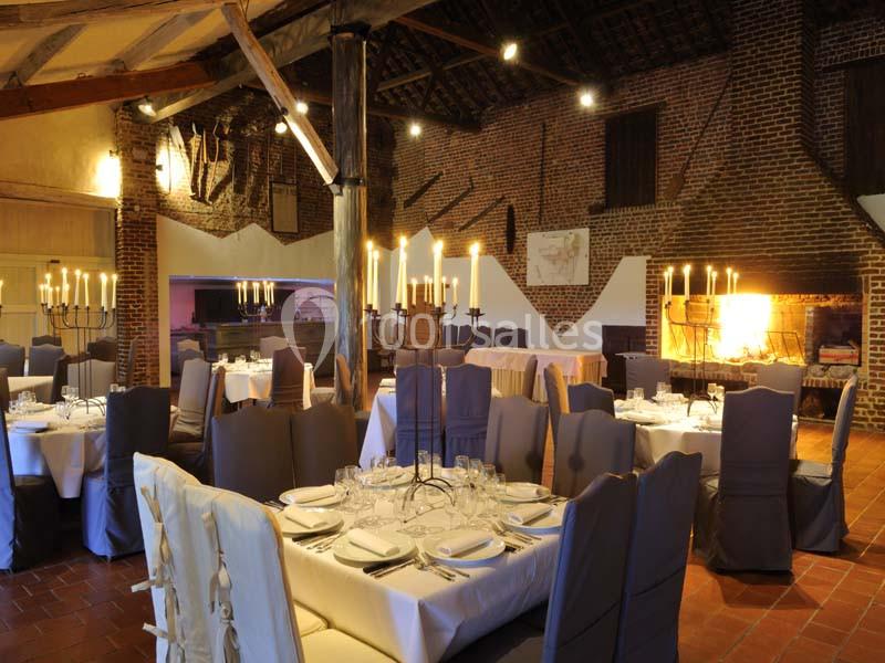 Salle de réception chaleureuse avec tables dressées, chaises couvertes et cheminée allumée dans un décor rustique.