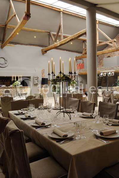 Salle de réception avec tables dressées, nappes beiges, chandeliers et poutres apparentes sous un toit vitré.