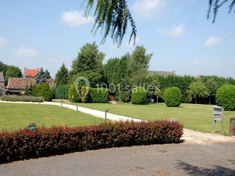 Allée bordée de pelouse et d'arbustes menant à des bâtiments en briques rouges, entourés de végétation.