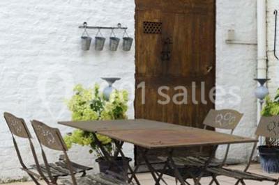 Miniature Location salle Pont-à-Celles (Hainaut) - La Ferme de la Pitance #12 Table et chaises en métal sur une terrasse en dalles, devant un mur blanc avec une porte en bois et des plantes décoratives.