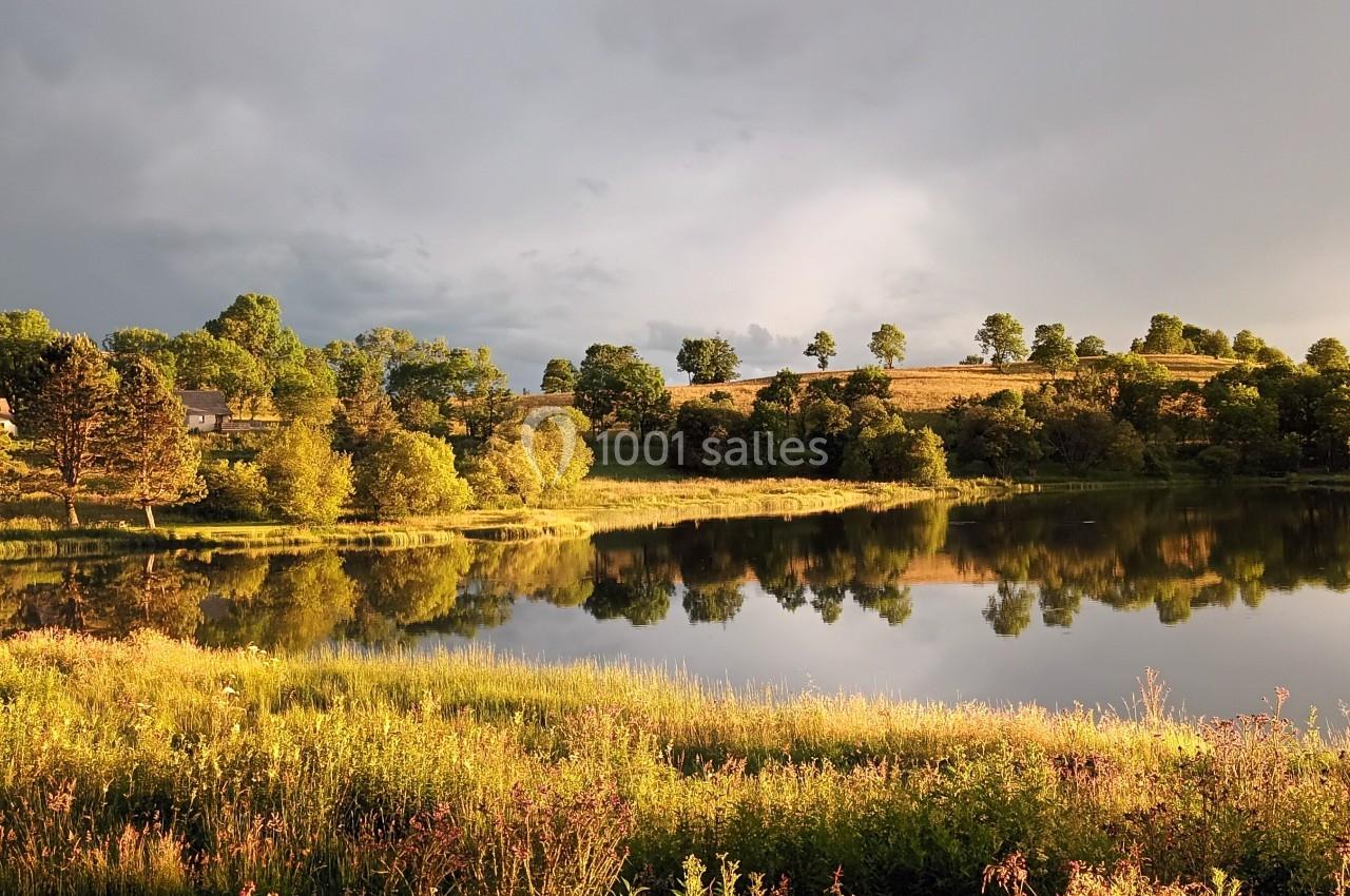 Paysage naturel avec un lac reflétant des arbres et des collines sous une lumière douce en fin de journée.