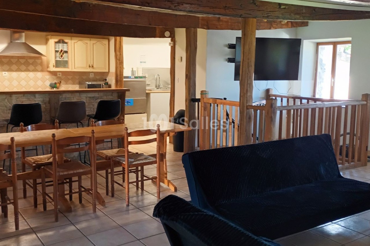 Salle à manger ouverte avec table en bois, cuisine équipée au fond, salon avec canapés et télévision à droite.
