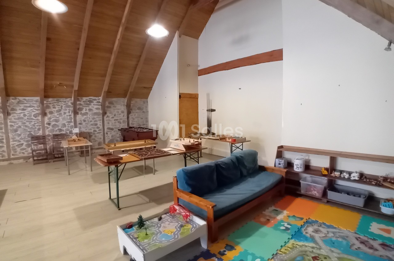 Salle avec un canapé, tapis coloré, tables en bois et jeux de société, sous un plafond en bois incliné.