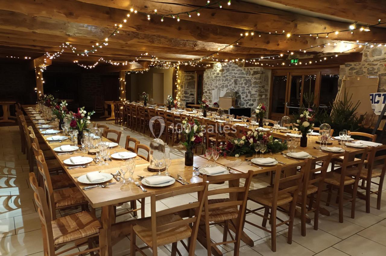 Salle décorée pour un repas, avec longues tables en bois, vaisselle dressée, guirlandes lumineuses et fleurs.
