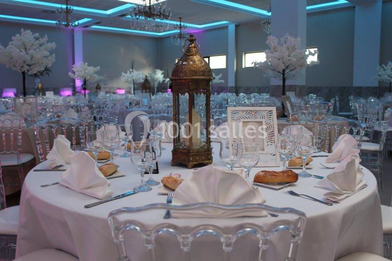 Table élégamment dressée avec nappes blanches, lanternes décoratives, vaisselle raffinée et arbres artificiels en arrière…