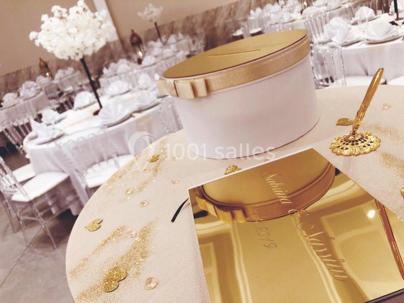 Boîte dorée pour cartes sur une table décorée, entourée de paillettes et d'une salle de réception élégante.