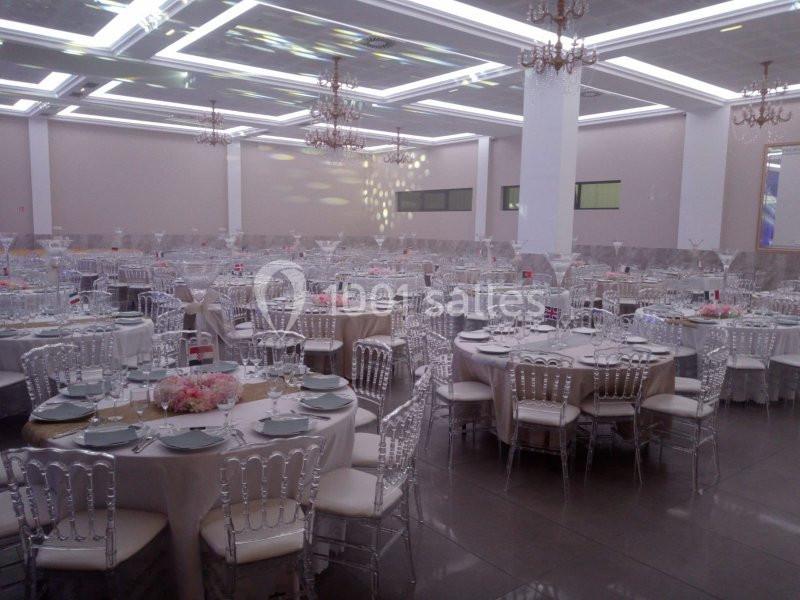 Salle de réception avec tables rondes dressées, nappes claires, chaises transparentes et décorations florales.