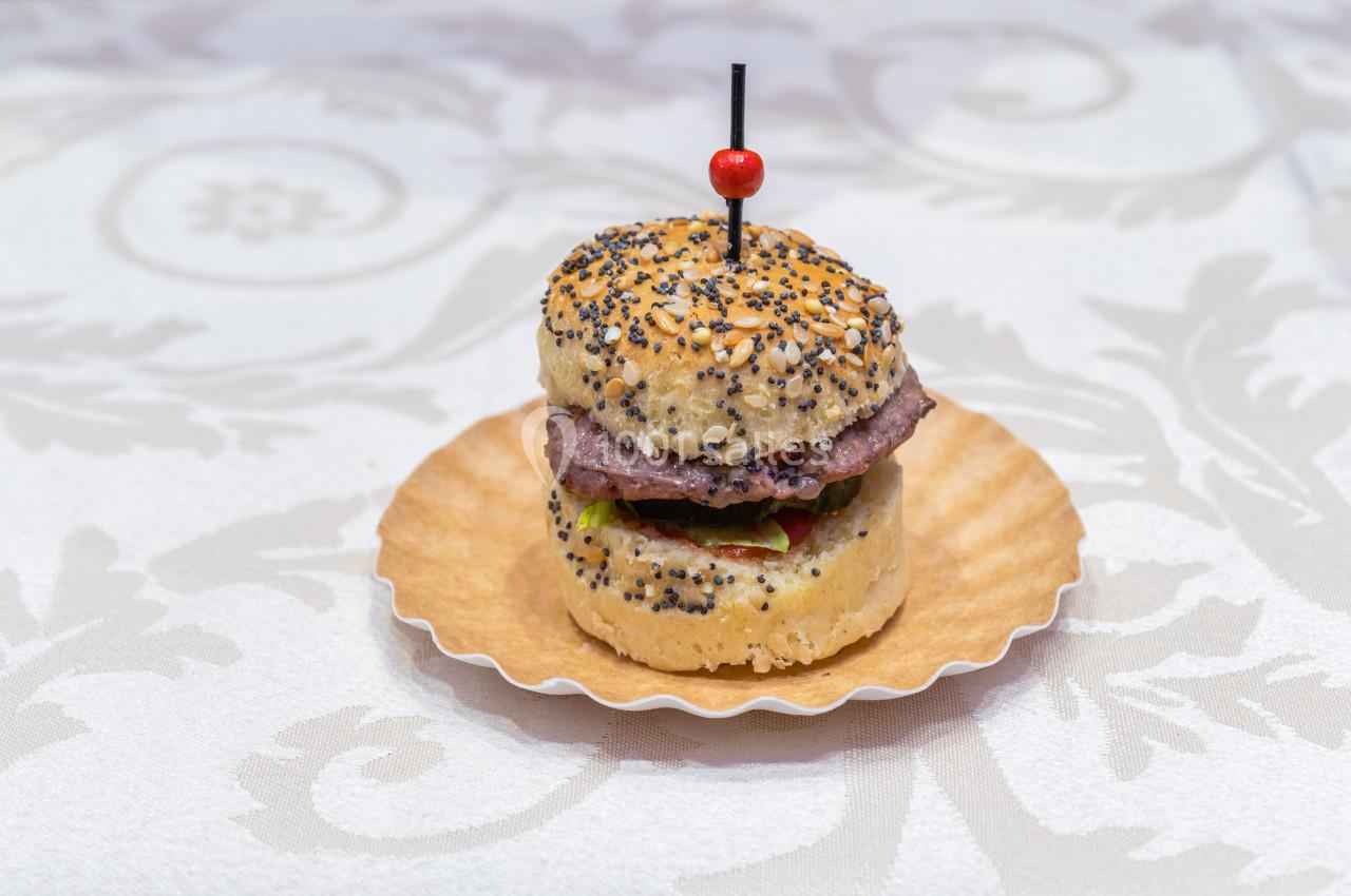 Mini burger sur un pain aux graines, garni de viande et de légumes, présenté sur une assiette en papier.