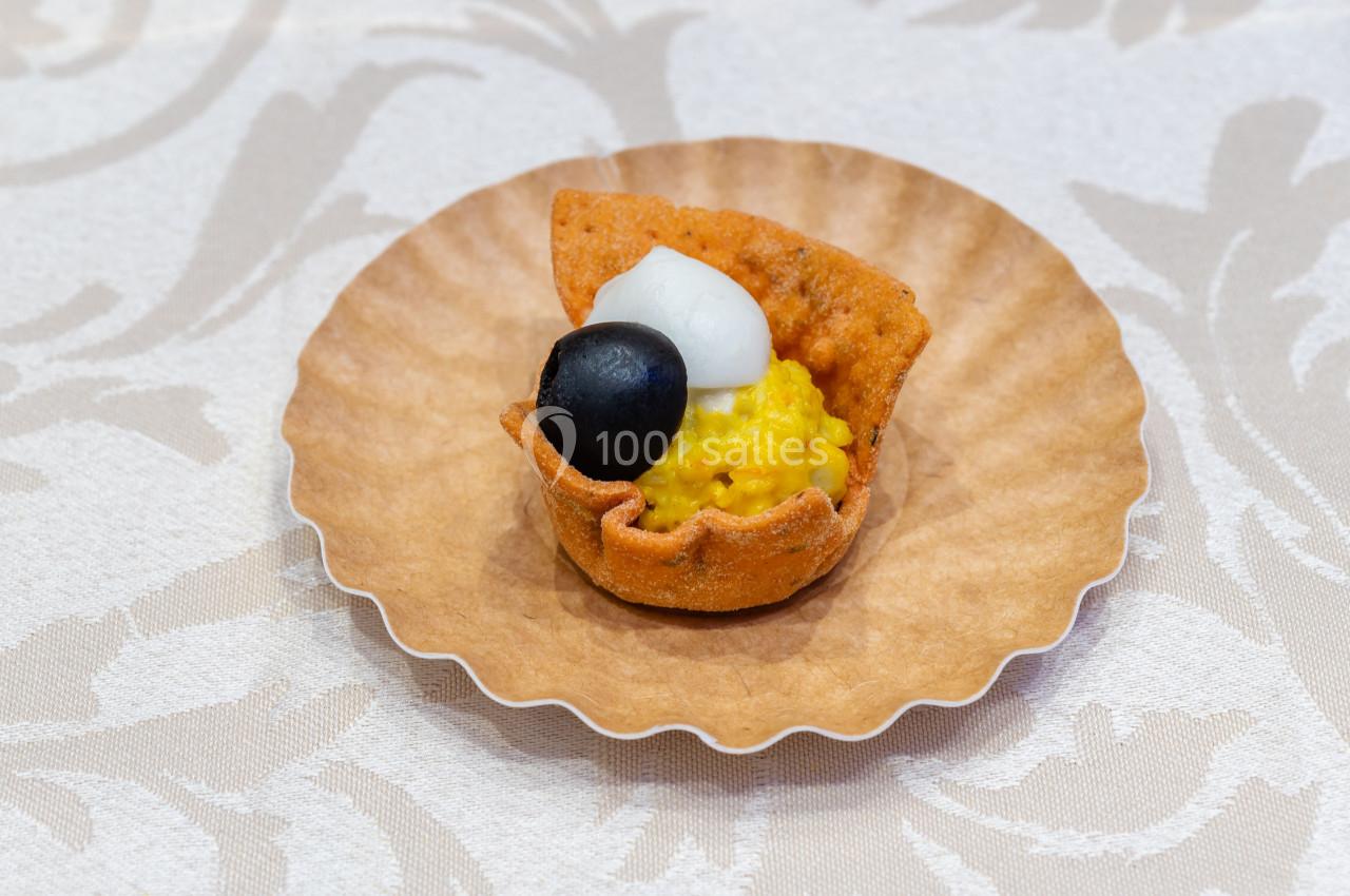 Tartelette apéritive garnie de maïs, d'une olive noire et d'une touche de crème blanche, présentée sur une assiette en…