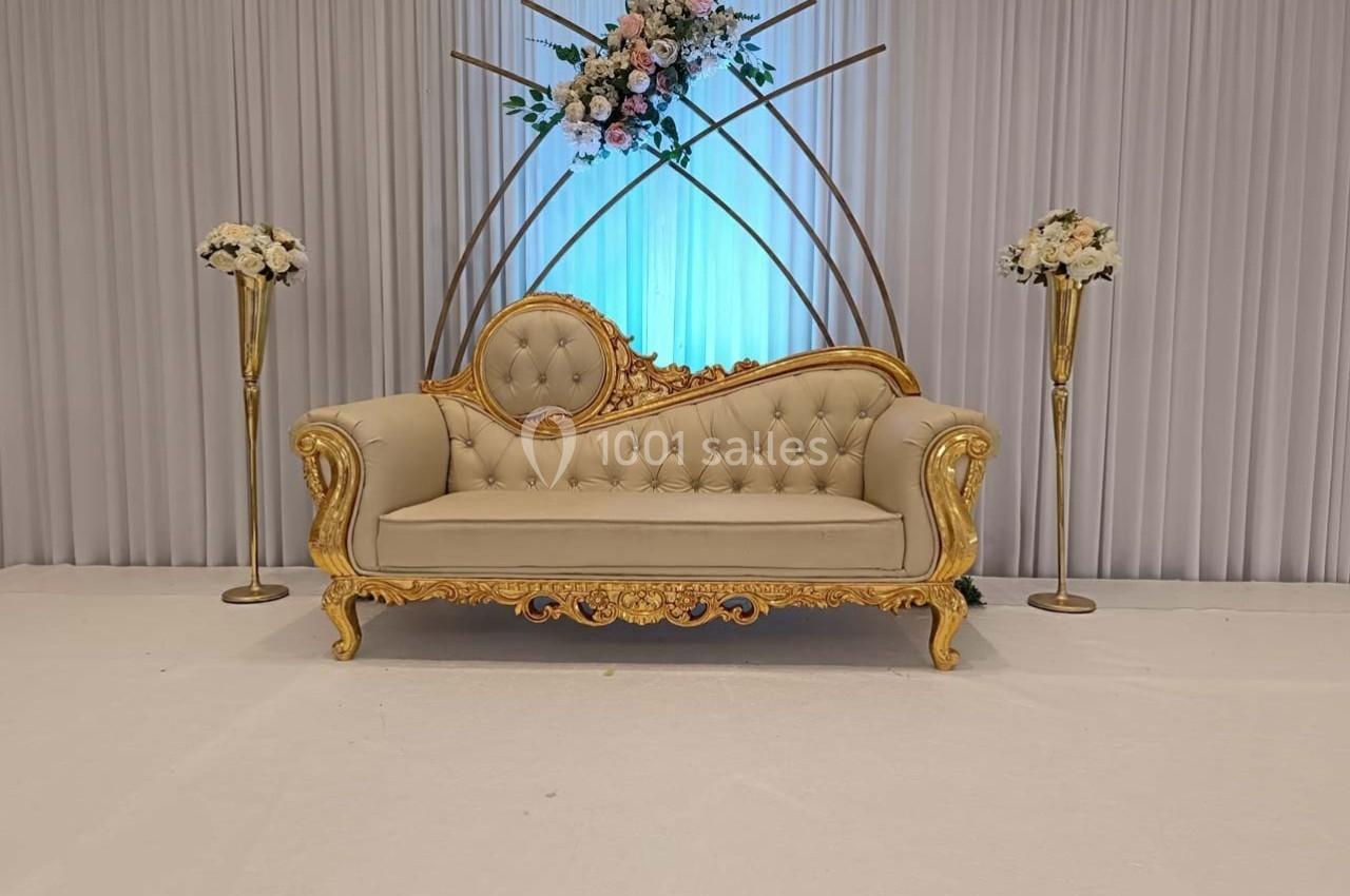 Canapé doré et capitonné devant un rideau blanc, décoré de fleurs et encadré par des supports floraux dorés.