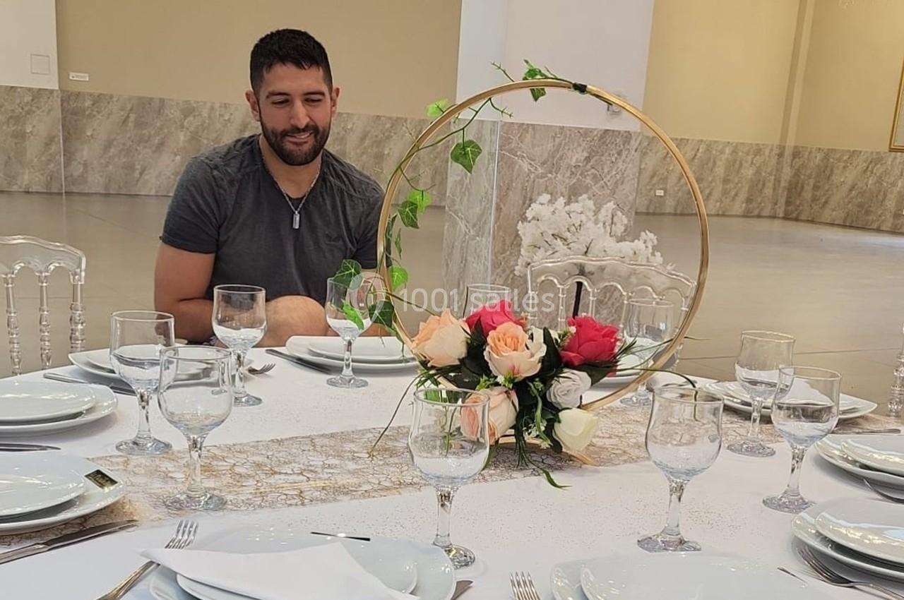 Un homme assis à une table élégamment dressée avec des fleurs et des verres, dans une grande salle lumineuse.
