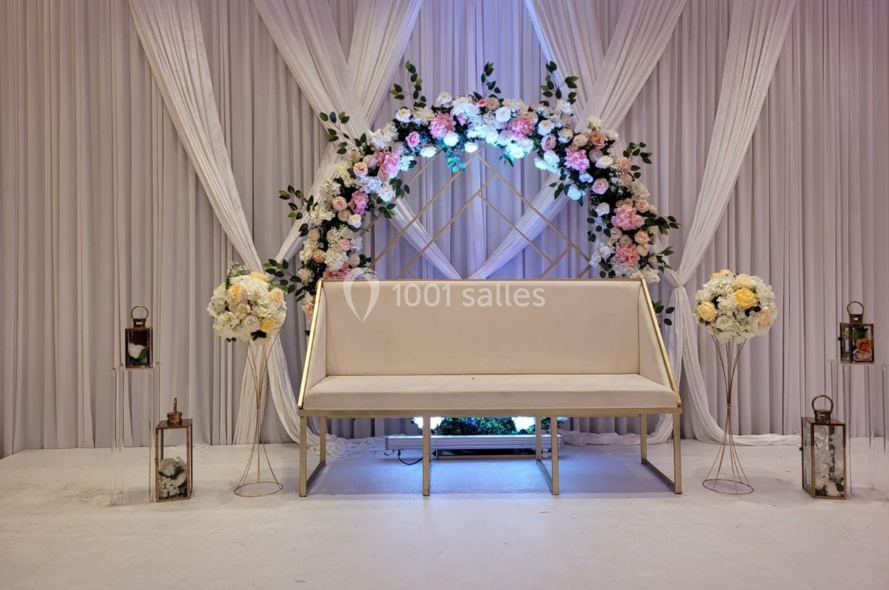 Banc beige devant une arche florale ornée de roses blanches et roses, avec des rideaux blancs en arrière-plan.