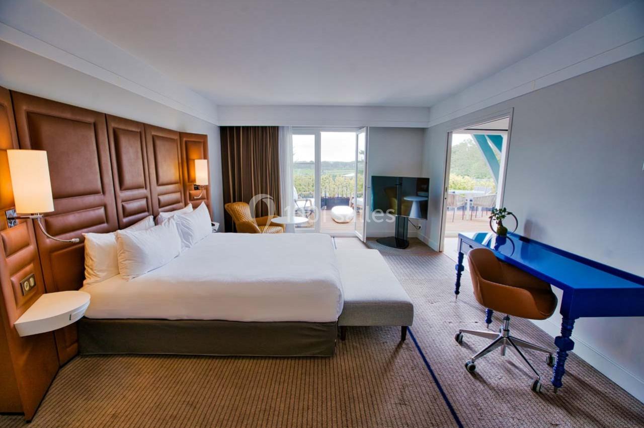 Location salle Rueil-Malmaison (Hauts-de-Seine) - Paris Country Club #33 Chambre d'hôtel lumineuse avec lit double, bureau bleu, fauteuil, télévision et balcon offrant une vue sur l'extérieur.