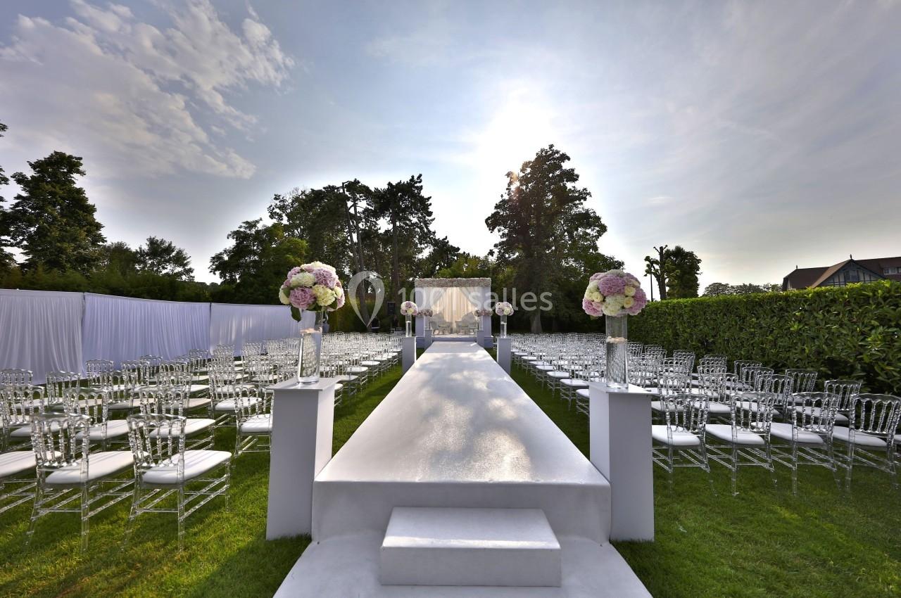 Location salle Rueil-Malmaison (Hauts-de-Seine) - Paris Country Club #11 Allée centrale blanche bordée de chaises transparentes et de bouquets, menant à une arche de mariage en extérieur.
