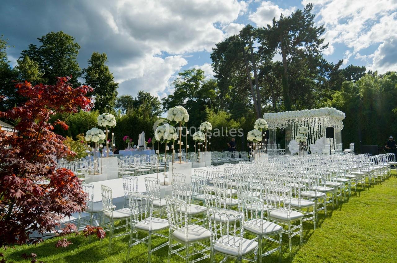 Location salle Rueil-Malmaison (Hauts-de-Seine) - Paris Country Club #12 Chaises transparentes disposées en rangées sur une pelouse, avec des décorations florales blanches et une arche en arrière…