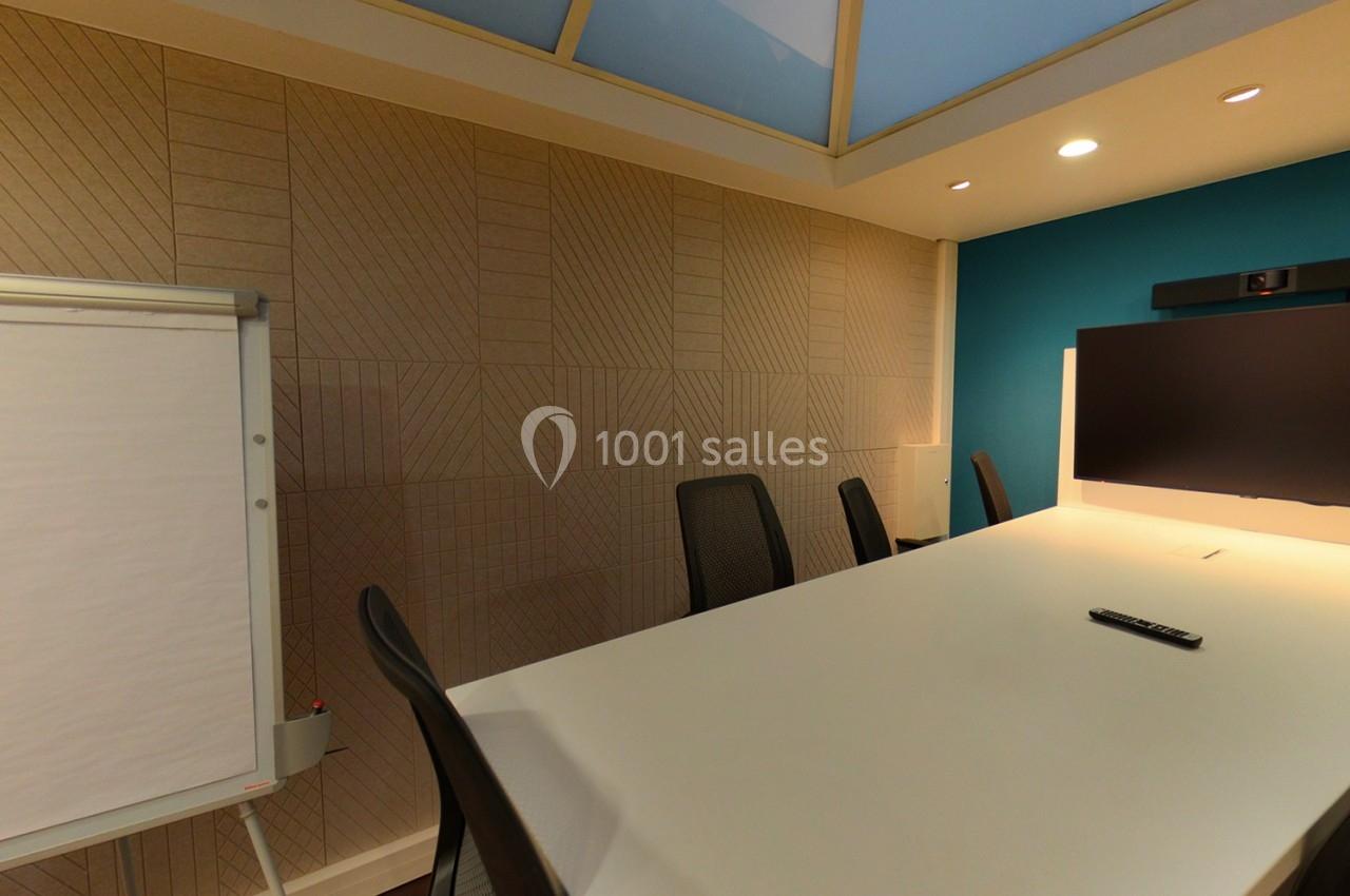 Salle de réunion moderne avec table blanche, chaises noires, écran mural, caméra de visioconférence et tableau blanc.
