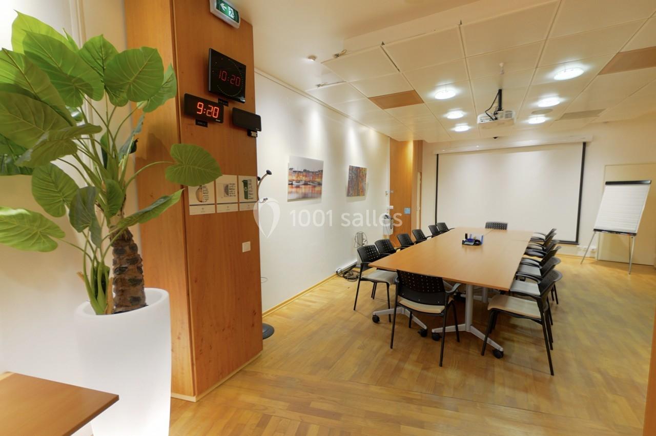 Salle de réunion lumineuse avec table rectangulaire, chaises, tableau blanc, horloge murale et plante décorative.