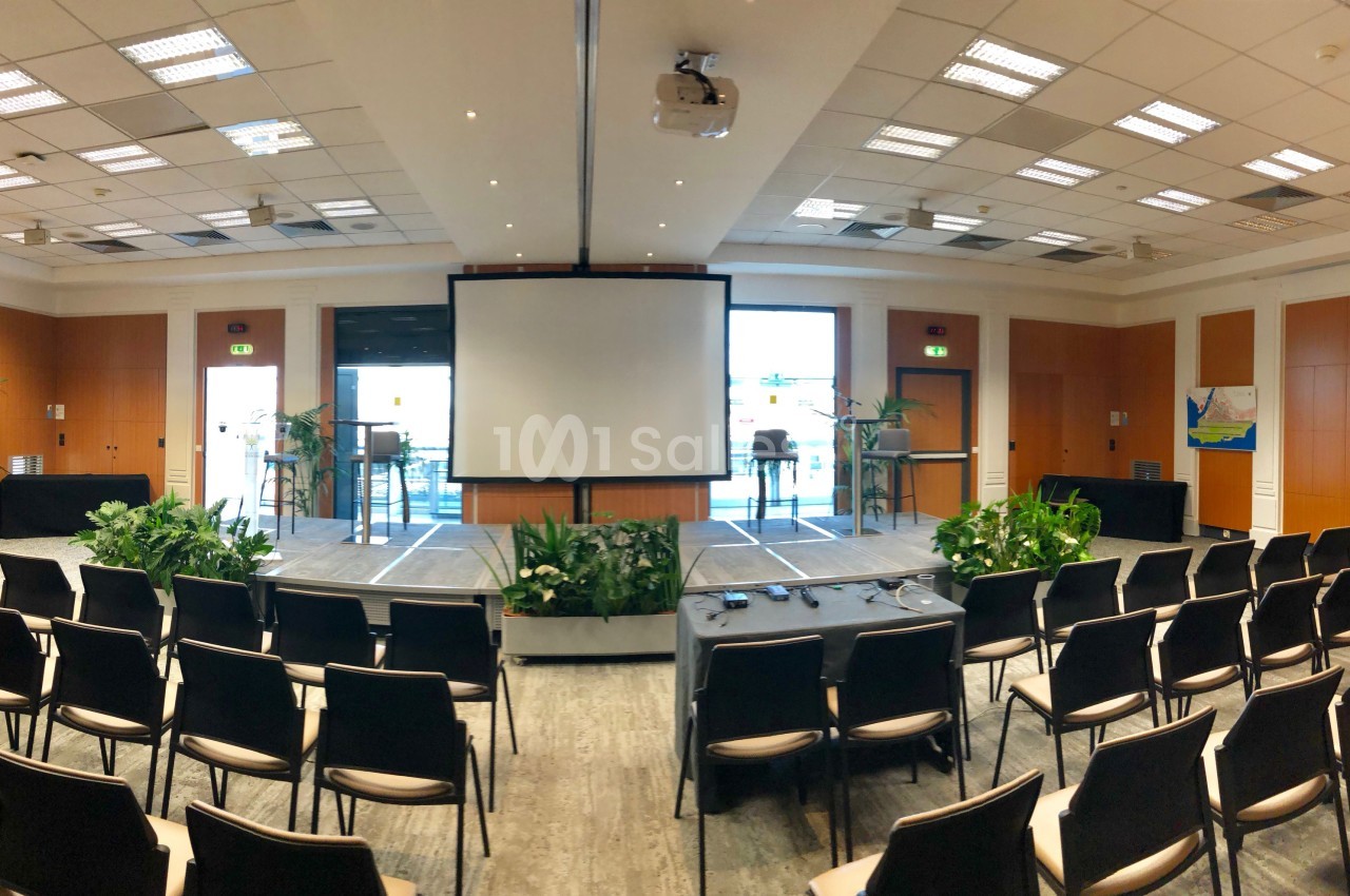 Salle de conférence lumineuse avec écran de projection, chaises alignées et plantes décoratives.