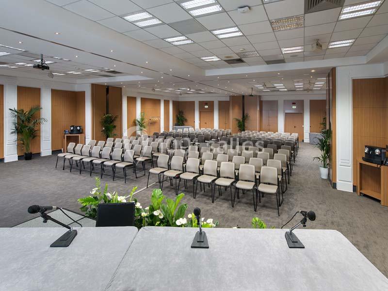 Salle de conférence moderne avec rangées de chaises vides, vue depuis une table équipée de micros.