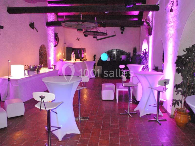 Salle décorée pour un événement, avec tables hautes, éclairage violet et mobilier moderne sur un sol en terre cuite.