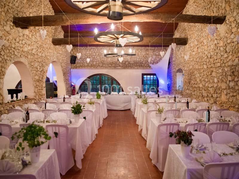 Salle de réception en pierre avec tables décorées de nappes blanches, éclairage chaleureux et poutres apparentes.