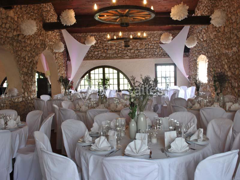 Salle de réception en pierre avec tables rondes dressées, nappes blanches et décorations suspendues.