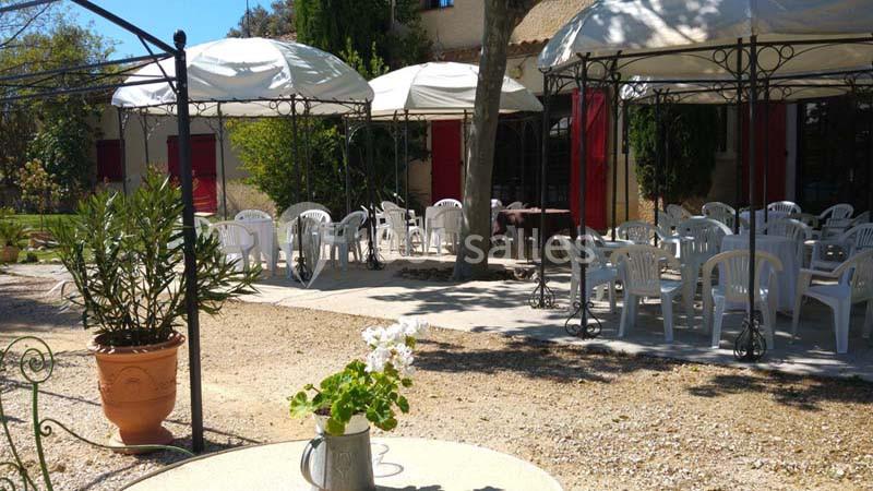 Terrasse extérieure avec tables et chaises blanches sous des parasols, entourée de végétation et de bâtiments en arrière…