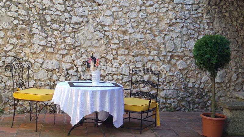 Table ronde avec nappe blanche, deux chaises jaunes et un petit arbre en pot devant un mur de pierres.