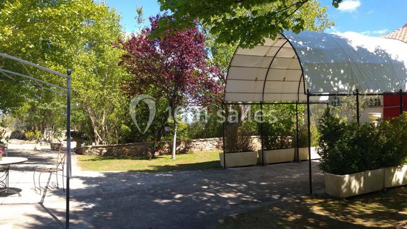 Abri en toile blanche dans un jardin arboré avec des tables en métal et des plantes en pot.