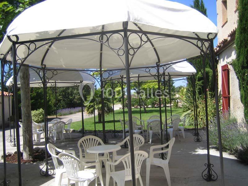 Des tonnelles blanches avec des tables et chaises en plastique blanc sur une terrasse, entourées de verdure.
