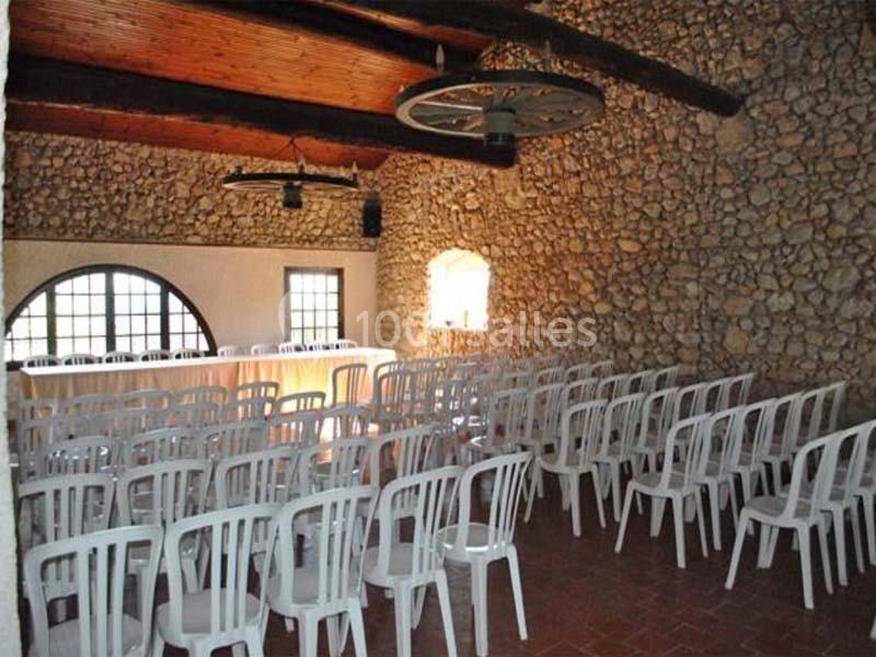 Salle en pierre avec des murs en pierre apparente, des poutres en bois au plafond et des chaises blanches alignées.