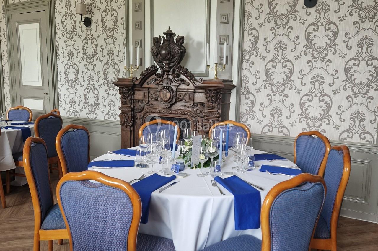 Salle élégante avec table ronde dressée, nappes blanches et bleues, chaises rembourrées, et cheminée décorative en arrière…