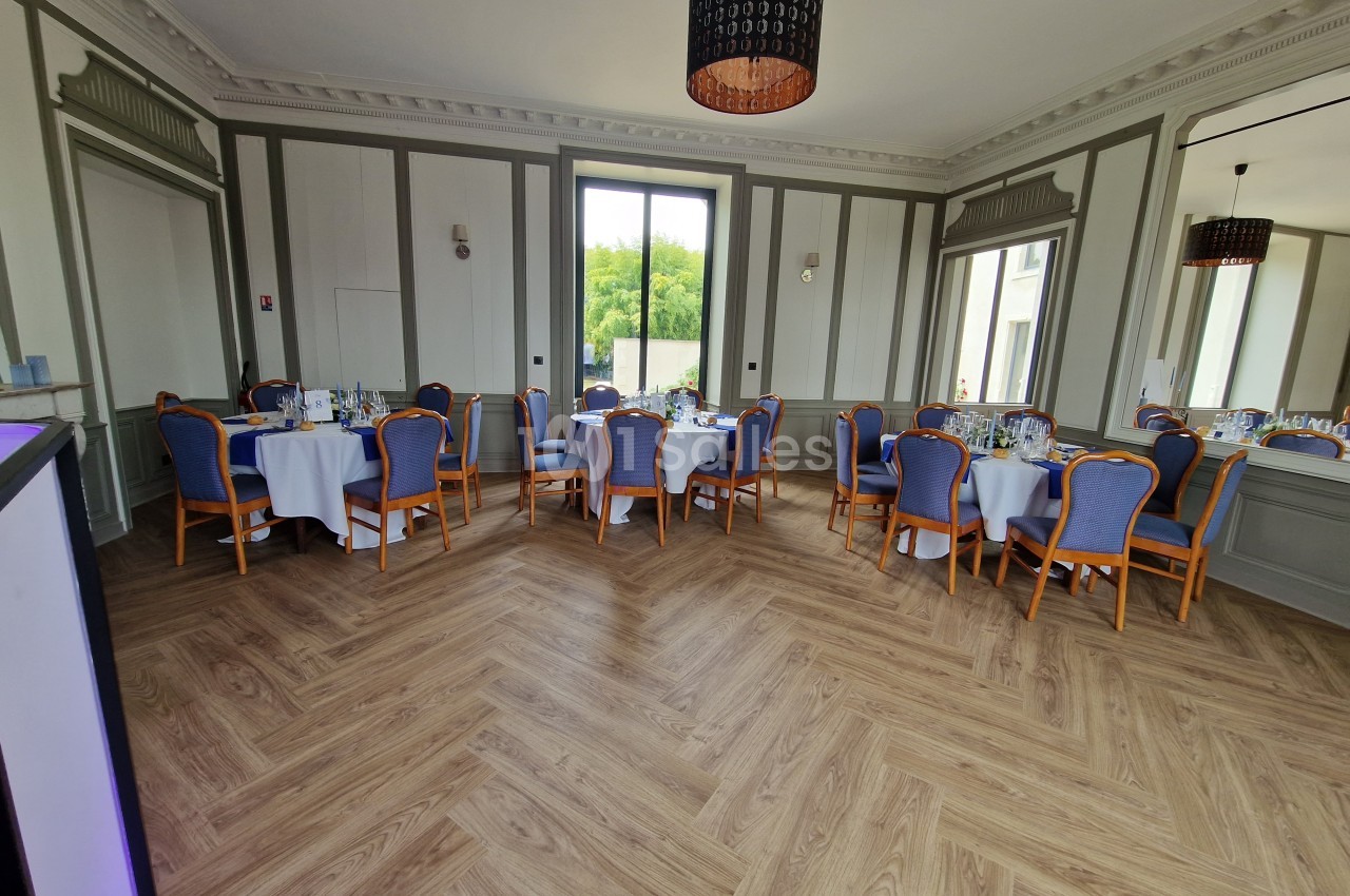 Salle élégante avec parquet en chevrons, tables rondes dressées et chaises bleues, éclairée par une grande fenêtre.