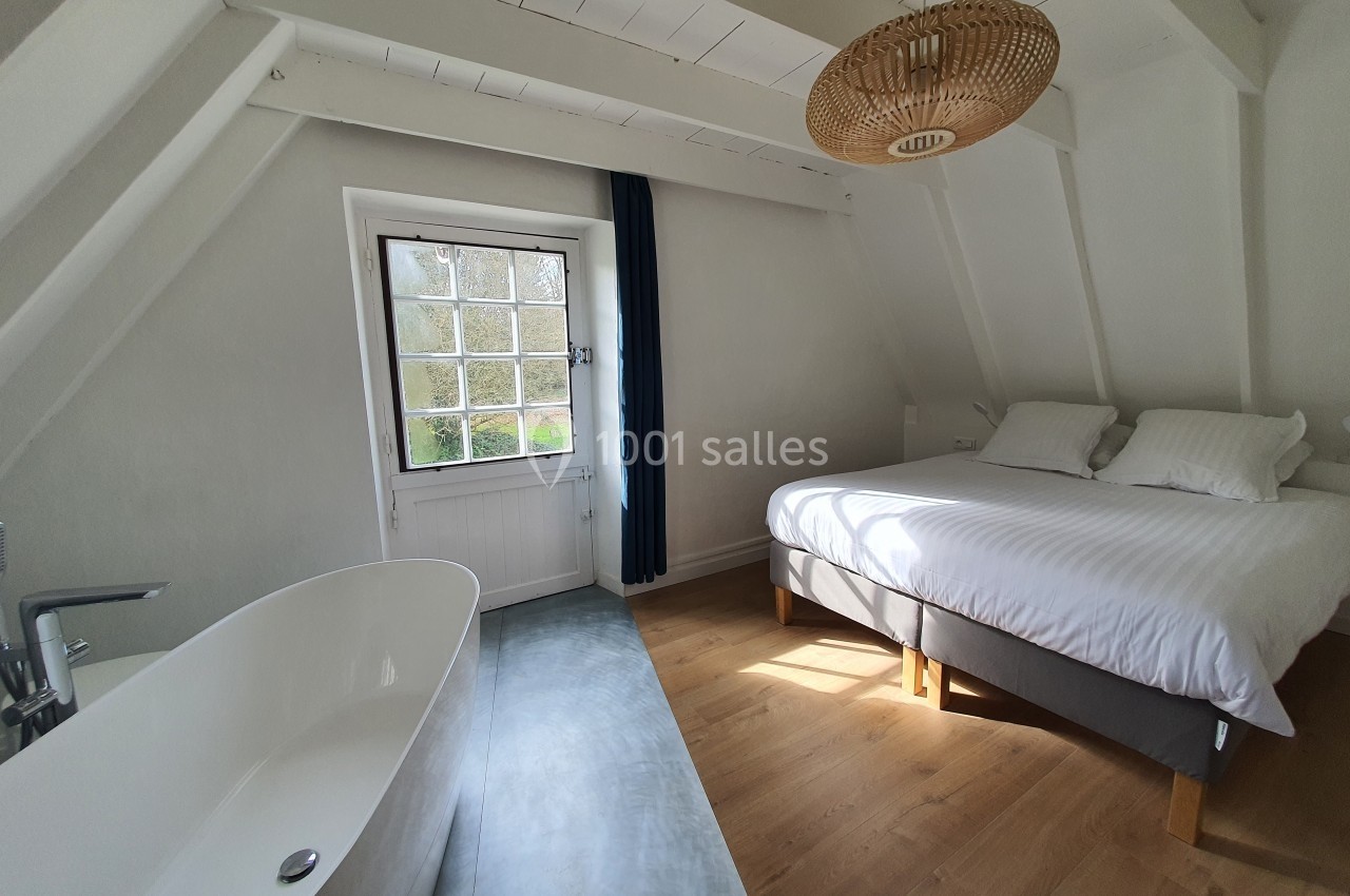 Chambre lumineuse avec lit double, baignoire blanche, parquet en bois et fenêtre donnant sur un paysage verdoyant.