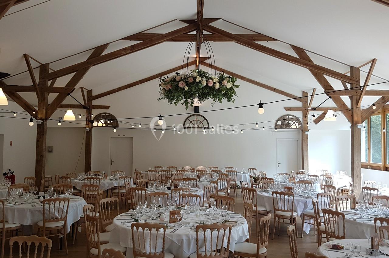 Salle de réception lumineuse avec tables rondes dressées, chaises en bois et décoration florale suspendue.
