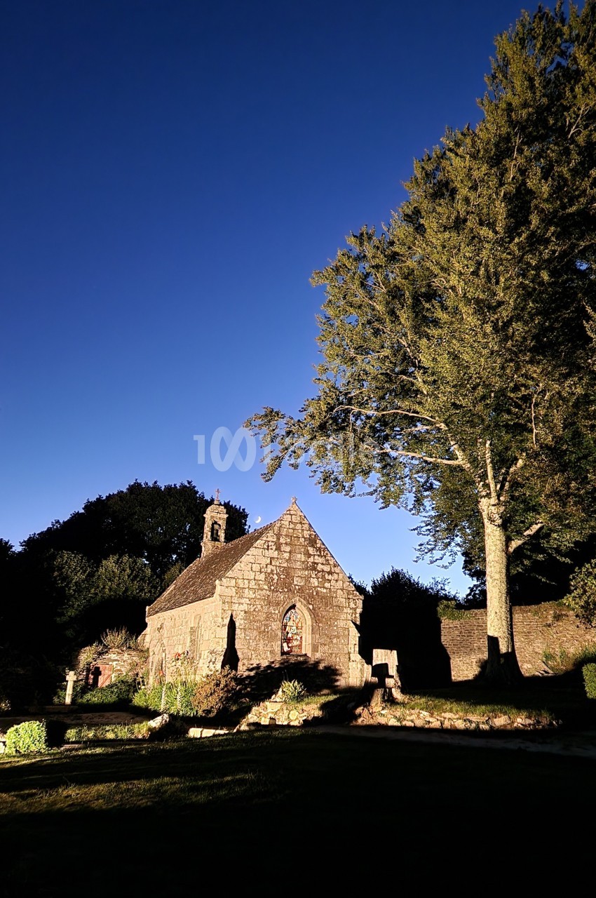 Une petite chapelle en pierre éclairée par une lumière chaude, entourée d'arbres sous un ciel bleu profond.